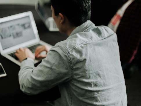 young man with black hair in a denim jacket with his back to the camera working at a laptop