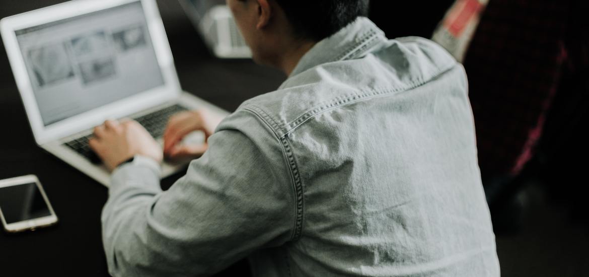 young man with black hair in a denim jacket with his back to the camera working at a laptop